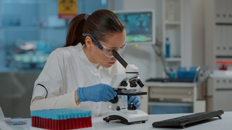 portrait of scientist looking through microscopic lens in laboratory, working on experiment with magnifying glass. woman using microscope and analyzing dna substance for development in lab.