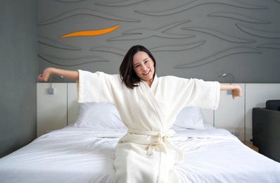 young asian woman in bathrobe rest on vacation, sitting in bed at home. stretch after get up in the morning