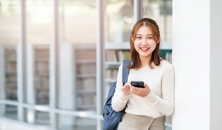 portrait of young beautiful university student with backpack holding smartphone, smiling and looking at camera, standing outside library room with copy space. back to school.