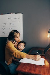 busy mom writing notes during a business phone call. working mom carrying her son on her lap while sitting in her home office. mother of one making business plans while working remotely.