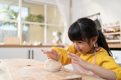 portrait of asian young kid girl doing homemade bakery in kitchen. adorable little child sitting on table feeling happy and enjoy learn to cooking foods or baking kneads yeast dough with hands at home