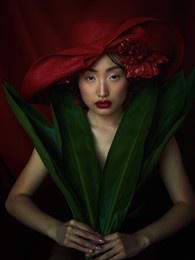 studio portrait of an asian girl on a dark background. the girl covers her body with large green leaves in a red hat                             