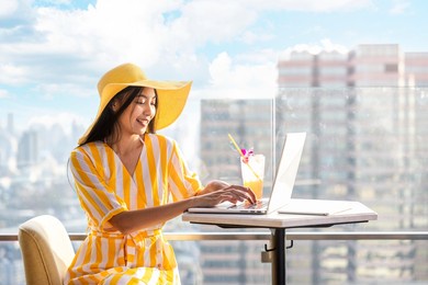 young chinese woman in summer yellow dress using laptop looking at computer screen enjoying watching funny social media video, sunny day. blurred background city scenery and beautiful sky and clouds.
