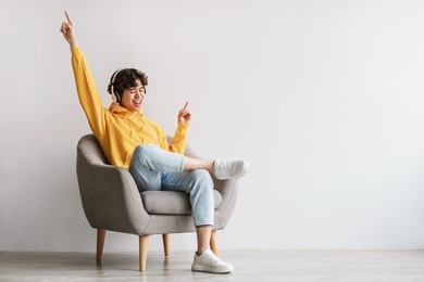 joyful young asian guy in wireless headphones sitting in armchair, having fun, listening to music, raising his arm, dancing to favorite song, enjoying cool soundtrack against white wall, empty space