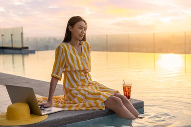 enjoy life. a elegant luxury woman in a yellow dress enjoys the summer sunset by the pool on skyscraper and laptop, hat, cocktail on edge of pool. scenic of orange sunset sky on blur background.  