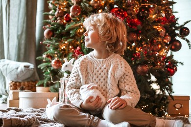little kid boy with blond hair and blue eyes in beautiful knitted clothes sits in living room near christmas tree in chalet with white small rabbits for family celebration of christmas and new year