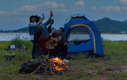 selective focus to bonfire. group of asian friends camping and drinking beverages with playing guitar and singing fun near tent and campfire an outdoors the evening with lake and mountain background.