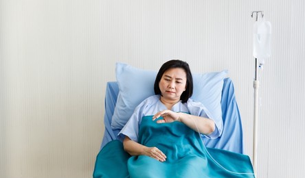asian senior stressed depressed upset doubtful sick female patient in blue hospital uniform laying down on bed alone looking at hand with injecting needle receiving normal saline solution from bag.