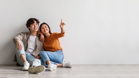 carefree millennial asian man and woman sitting on floor near white studio wall and pointing at copy space, banner design. young couple offering place for cool advertisement, panorama