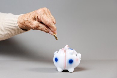 wrinkled hand of an elderly woman puts a euro coin in a piggy bank. gray background. the concept of savings and banking.