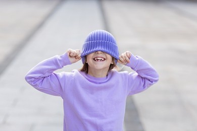 cute little caucasian girl eight years old with blonde hair smiling outdoor. kid wearing stylish shirt and knitted hat violet color. trendy color of the 2022 year very peri