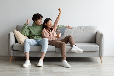 excited young asian couple sitting on couch with tablet pc, celebrating online win, great deal or business success at home, free space. millennial spouses enjoying big sale in web store