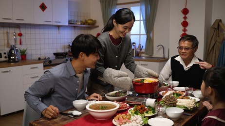 smiling asian daughter in law carrying and putting hot pot on portable stove. happy senior father gesturing to sit down at dining table, ready to start reunion meal