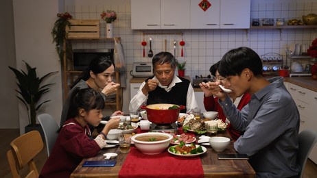 asian family members enjoying tasty soup together with spoons from the pot while having reunion dinner in the dining room at home on chinese new year's eve