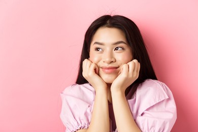 close-up of excited asian girl listening with interest, smiling amused and looking aside at logo, daydreaming, standing against pink background