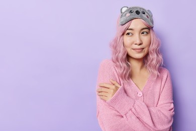 thoughtful asian woman with pink hair embraces herself wears pink soft jumper and sleepmask looks away with deamy expression isolated over purple background blank copy space for your promotion