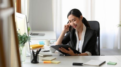 asian female manager talking on the phone with business client while looking at the informations on clipboard paper at her office workplace.