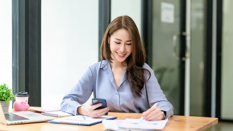 smiling beautiful asian businesswoman analyzing chart and graph showing changes on the market and holding smartphone at office.