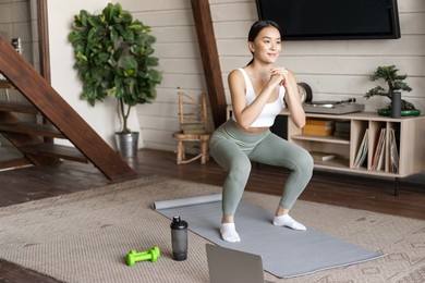 young asian fitness girl doing squats workout at home, follow online sport gym instructor, standing on floor mat and smiling, exercising in living room