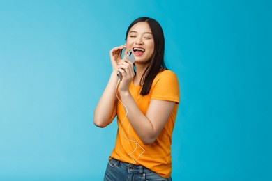 joyful carefree attractive asian woman close eyes pleasure and joy, smiling wear earphones, listening music via headphones, hold smartphone like microphone karaoke, singing along favorite song