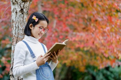 smiling asian cute little girl with glasses reading book under maple tree