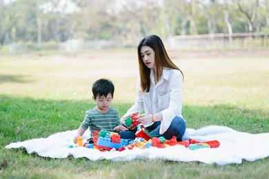 young asian mom and little son sitting on grass playing with educational toy in spend sunny day in summer park. family on busy face spend time together having fun with colorful blocks