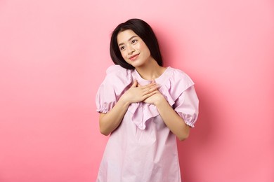 dreamy asian girl smiling and holding hands on chest, looking left at something romantic and cute, standing on pink background