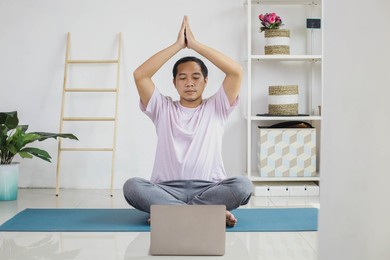 handsome young asian man sitting cross-legged on mat doing yoga while looking at laptop
