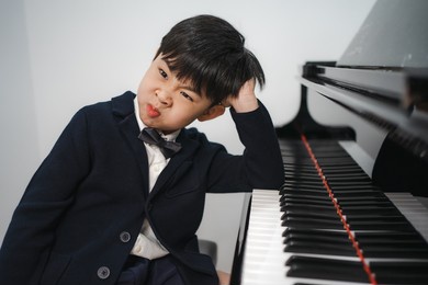 lazy little boy in black suit sitting at grand piano makes funny face. cute boring kid pianist feels tired to practice playing classic keyboard. playful child musician refuses to perform a concert.