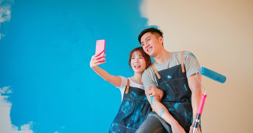 young asian couple decorate home - they take selfie by mobile phone after painting wall with blue color