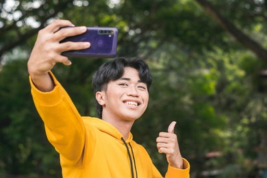 a young man in a yellow hoodie sweater takes a selfie while making a thumbs up sign. outdoor scene.