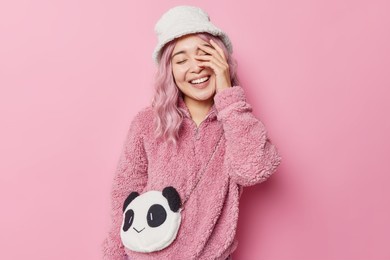 joyful young asian woman covers half of face with hand smiles happily keeps eyes closed smiles pleasantly feels pleased wears winter hat and coat isolated over pink background. emotions concept