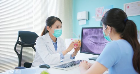asian female dentist showing teeth xray at dental clinic - she teaches patient for teeth brushing and tooth decay by denture model while they wear protective face mask to prevent covid19