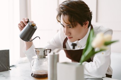 portrait of young asian barista man wearing brown apron preparing coffee using pour over coffee maker and drip in minimal cafe shop. alternative ways of brewing coffee. man barista cafe owner concept