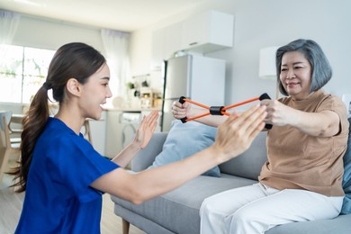 asian mature older woman doing physiotherapist with support from nurse. senior elderly female patient sitting on sofa in living room use resistance stretch band exercise with caregiver in nursing care