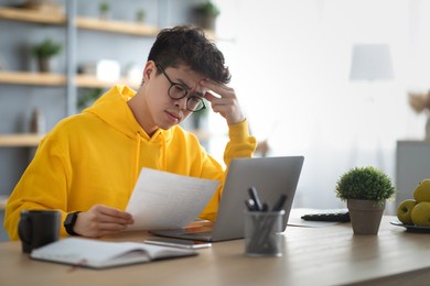 upset young asian man in eyeglasses sitting at table with modern laptop at home office, reading letter mail and touching his forehead, copy space. sad guy holding paper document or bills, got bad news