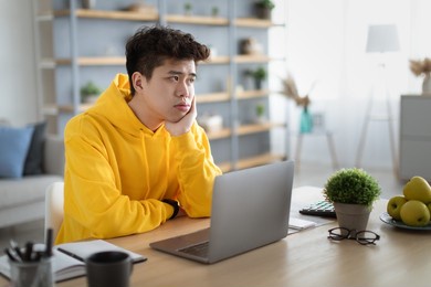 bored young asian guy sitting at desk using pc laptop, looking aside, thinking about something during online lesson or remote work at home office. tired man having dull distant job, feeling lazy