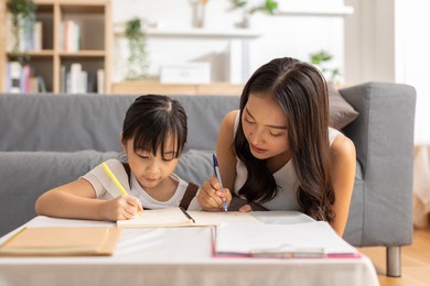 happy asian little girl doing homework with mother at home. asian young mother teaching small daughter to drawing reading and writing to develop her daughter skill. home school concept