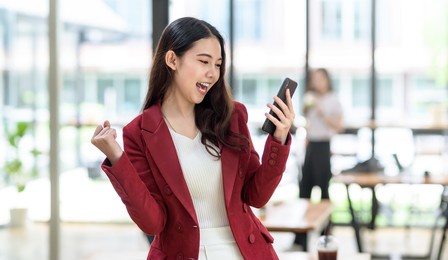 happy young asian business woman successful excited raised hands rejoicing with smartphone.