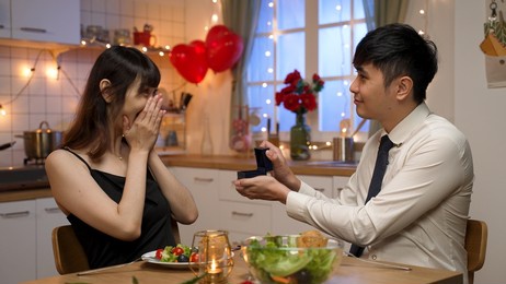 asian man wearing shirt and tie showing jewellery as valentine’s day present while his surprised girlfriend covering face and nodding head saying she likes it at dinner date at home