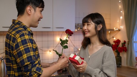 asian boyfriend showing surprise gift and red rose to his happy girlfriend on valentine’s day at decorated cozy home interior