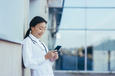 happy female doctor, smiling and rejoicing, asian woman uses the phone, during a break near the clinic medical apps, looking at screen, working online, chatting, consulting patient concept