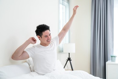 asian handsome male in pajamas wake up in the morning with happiness. attractive young man smiling, feeling happy and relax then stretching body after getting up from sleep on bed in bedroom at home.