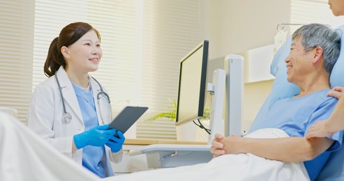 asian female doctor holding digital tablet talking to senior man lying in bed at hospital