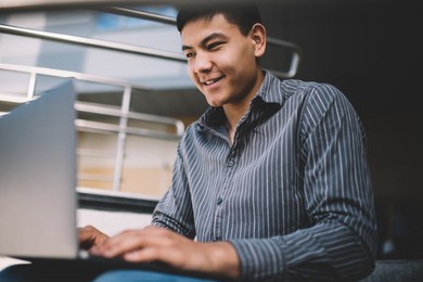 asian millennial guy watching on laptop while sitting on stairs near office building. concept of freelance and remote work. idea of modern successful man. young smiling male person. sunny day