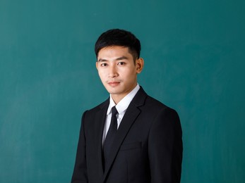 portrait studio shot of asian young professional successful male teacher in black formal suit and necktie standing smiling look at camera in front green blackboard background in classroom at school.