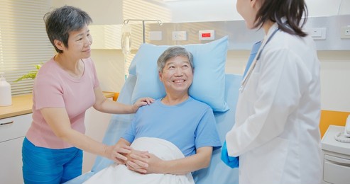 senior people healthcare concept - asian female doctor is talking to senior man patient lying in bed while elder woman take care of him at hospital
