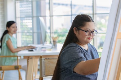 portrait shot of asian happy lovely mother standing smiling with young chubby down syndrome autistic autism little daughter wearing eyeglasses holding paintbrush drawing painting on canvas easel.