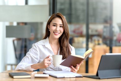 beautiful young asian businesswoman drinking a coffee and holding a book working on laptop at office. looking at camera.