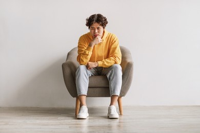 negative emotions. young asian man sitting in armchair with thoughtful face expression, feeling sad or tired, having problem, suffering from depression or stress against white studio wall